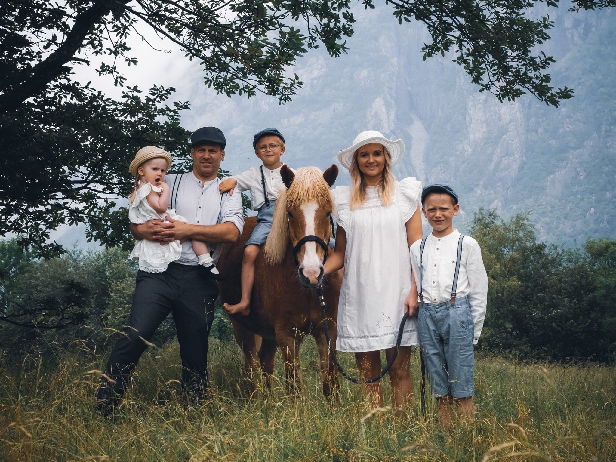 En familie på fem kledd i historiske klær. De står i gresset ved et tre sammen med en hest.