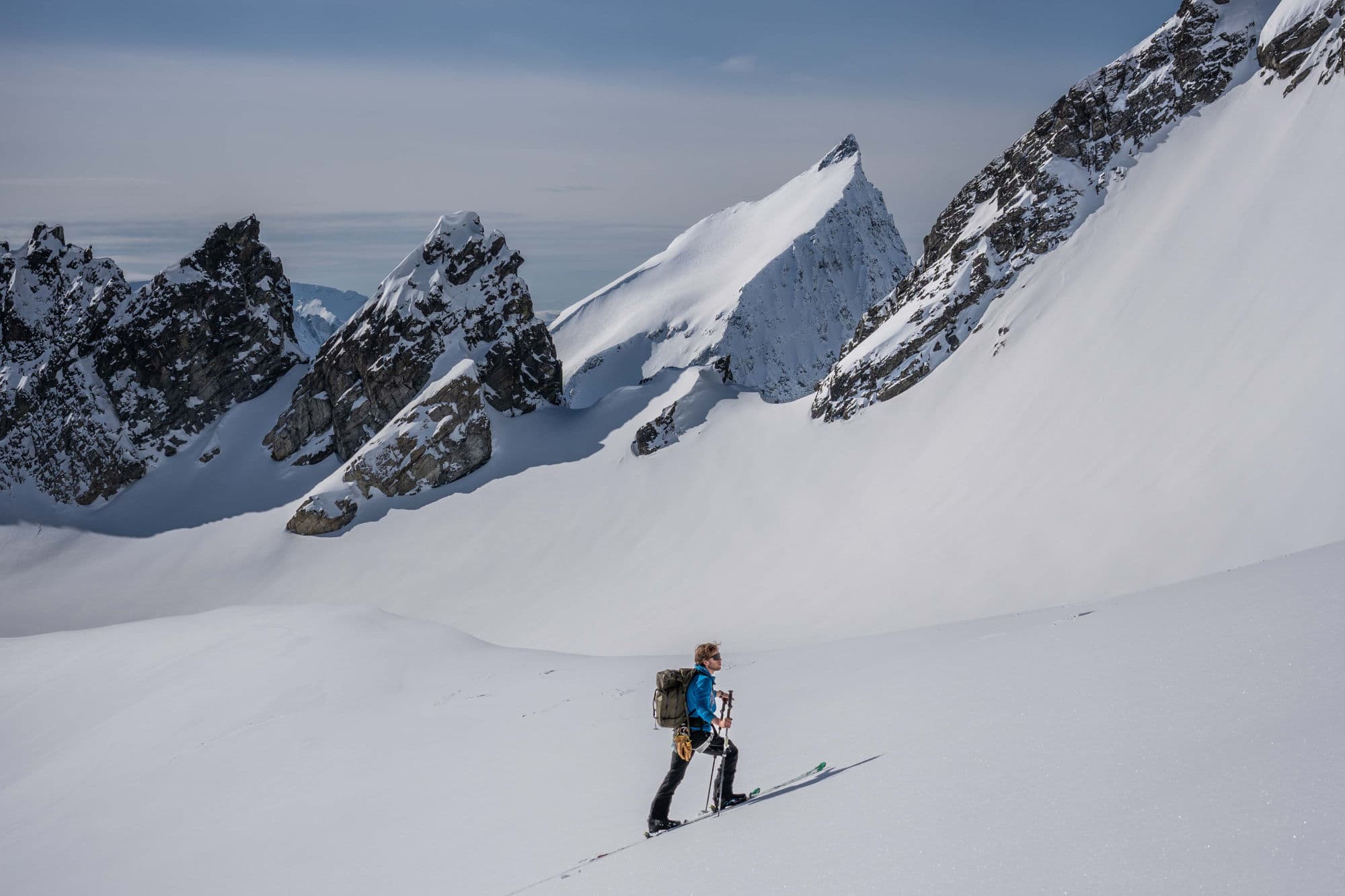 Person med ski på vei opp hvitkledde fjellsider, men Sunnmørsalpene og de taggede fjelltoppene i bakgrunnen.