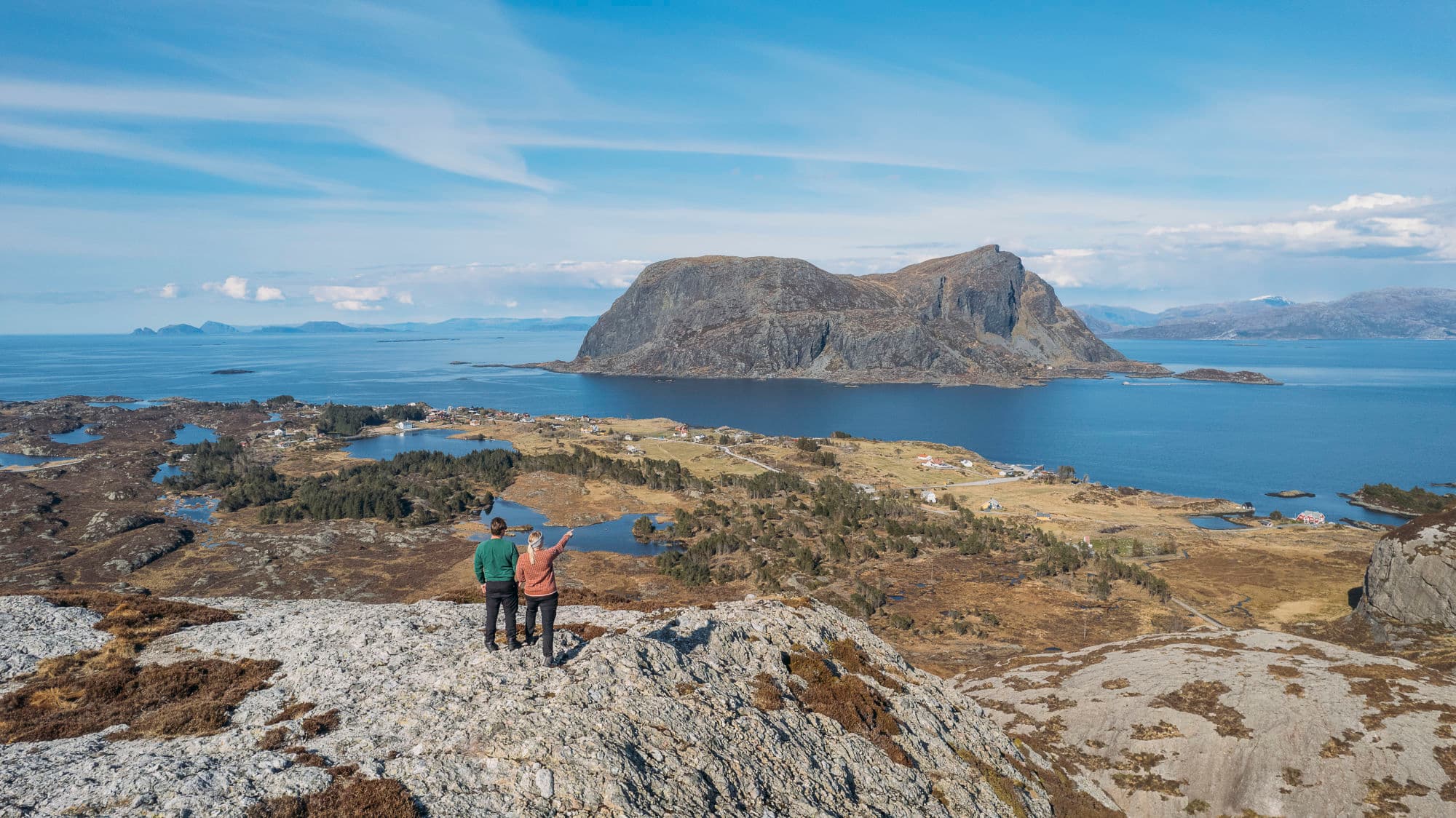 to personer står på toppen av eit fjell. eine personen peiker utover. utsikta viser mot et anna stort fjell som står plassert midt i sjøen. det er blått hav rundt, grøn, gul og oransje nyanser i landskapet, og grått fjell.
