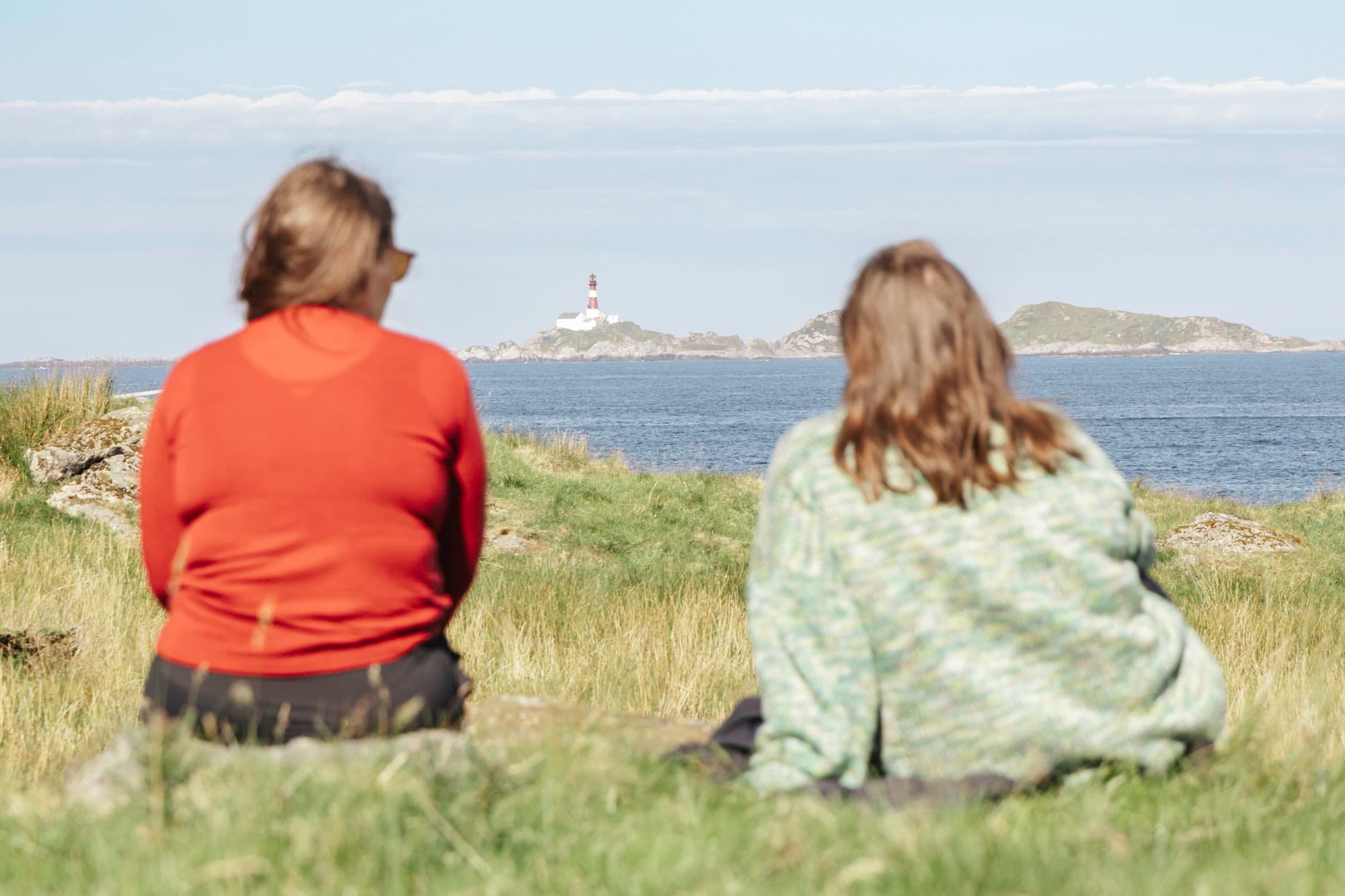 to jenter sitter på det grønne gresset og ser ut over sjøen. i sikte er eit fyrtårn på ei anna øy. det er blå himmel og blå sjø. jentene har på seg gensere, ho ein har raud genser medan ho andre har grøn genser
