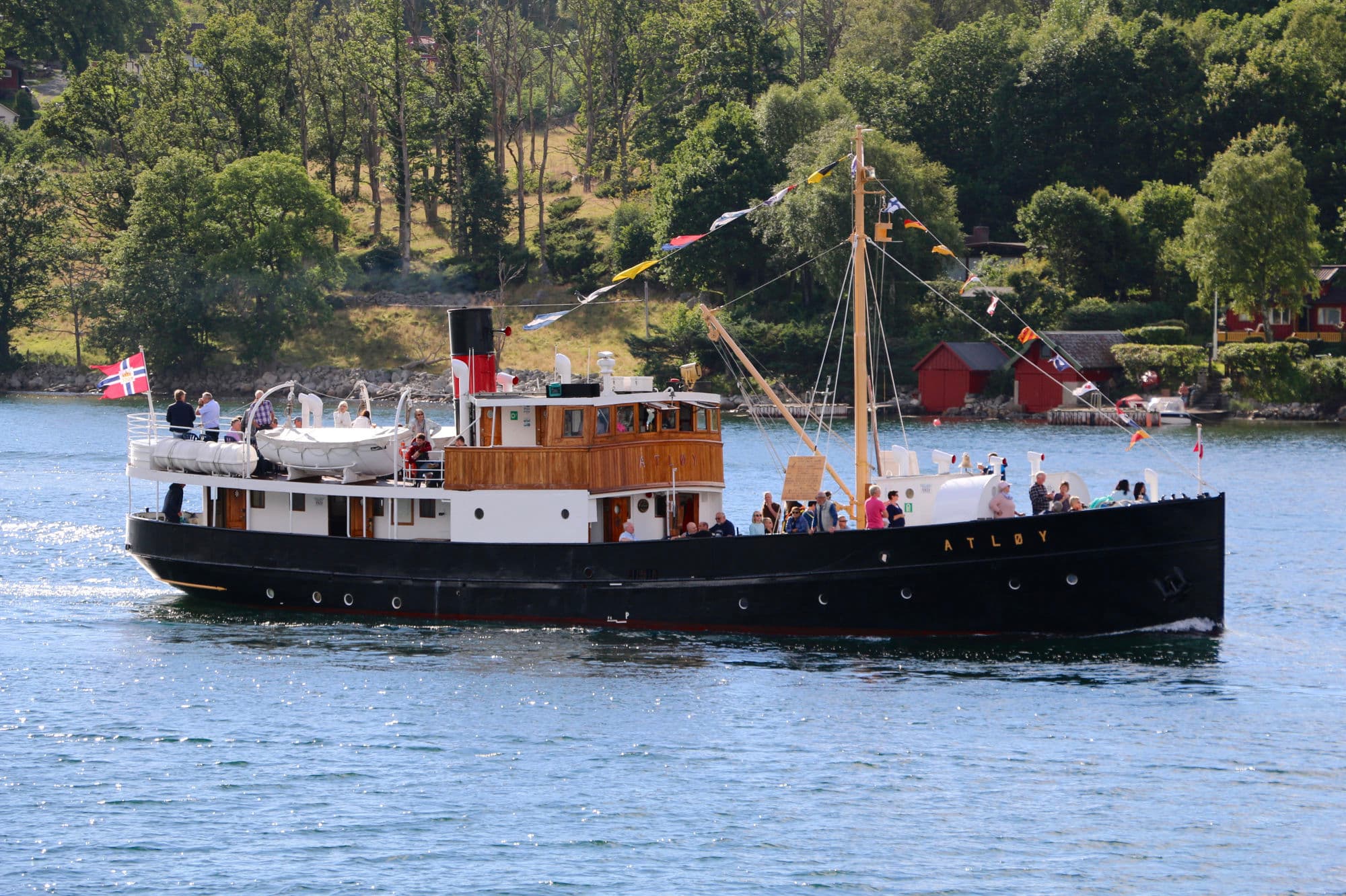 en veteranbåt med masse folk på. det er blått hav og grøne trær rundt