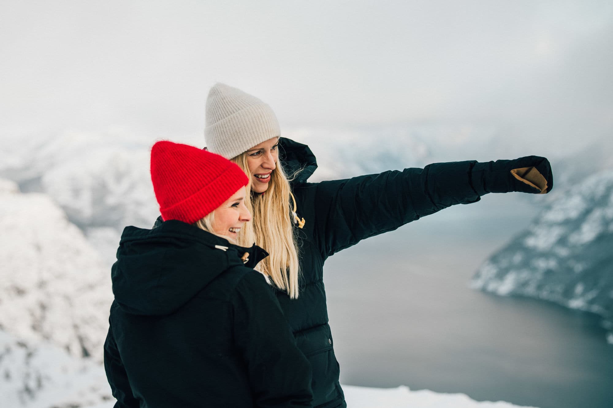 To damer i vinterklær peker på noe. Fjord og snødekte fjell rundt.