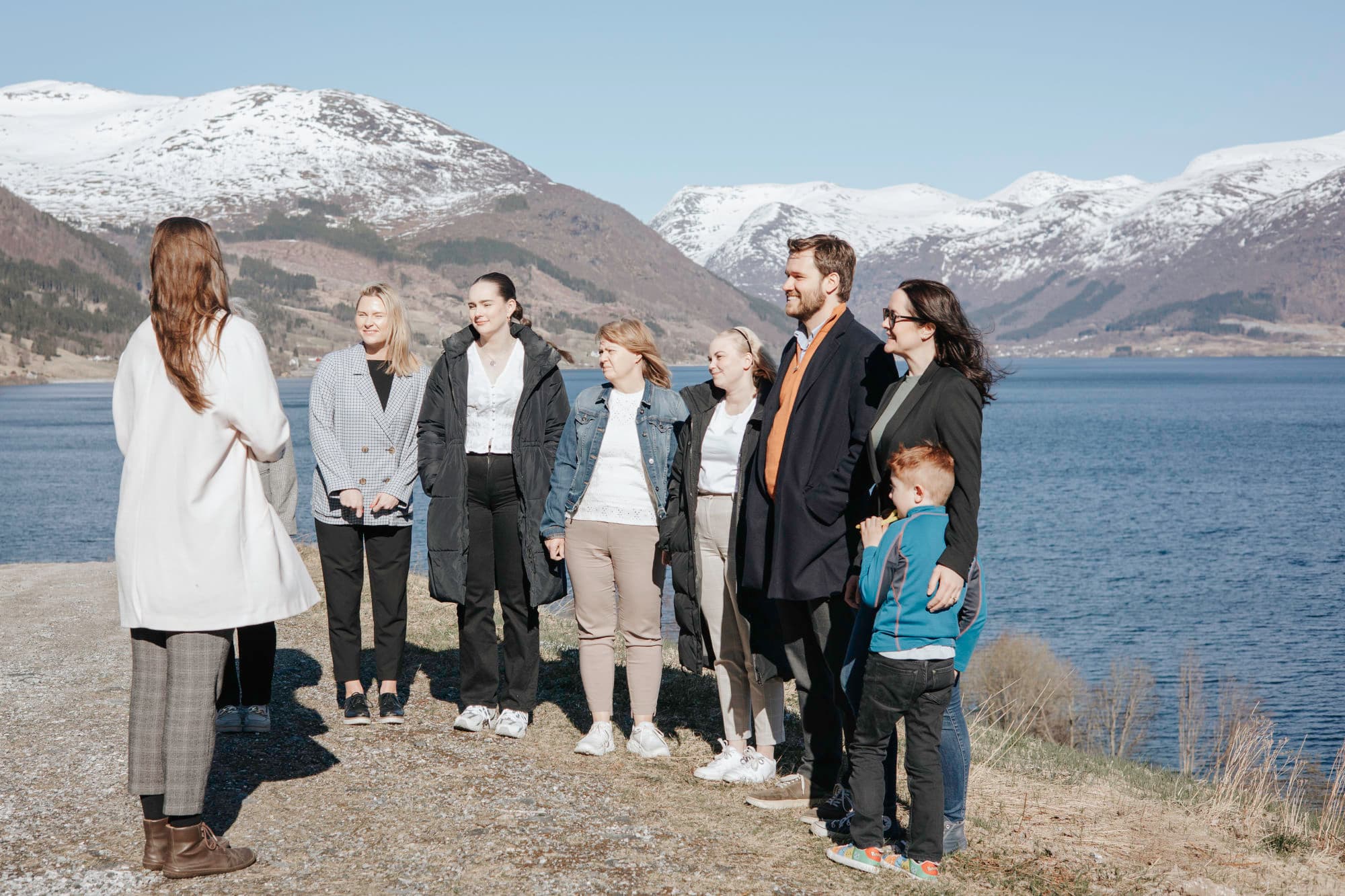 ein gruppe står og ser på ei dame som står og snakker. dei smiler og er interessert i kva ho snakker om. bak gruppen er det utsikt over høge fjell og blått vatn