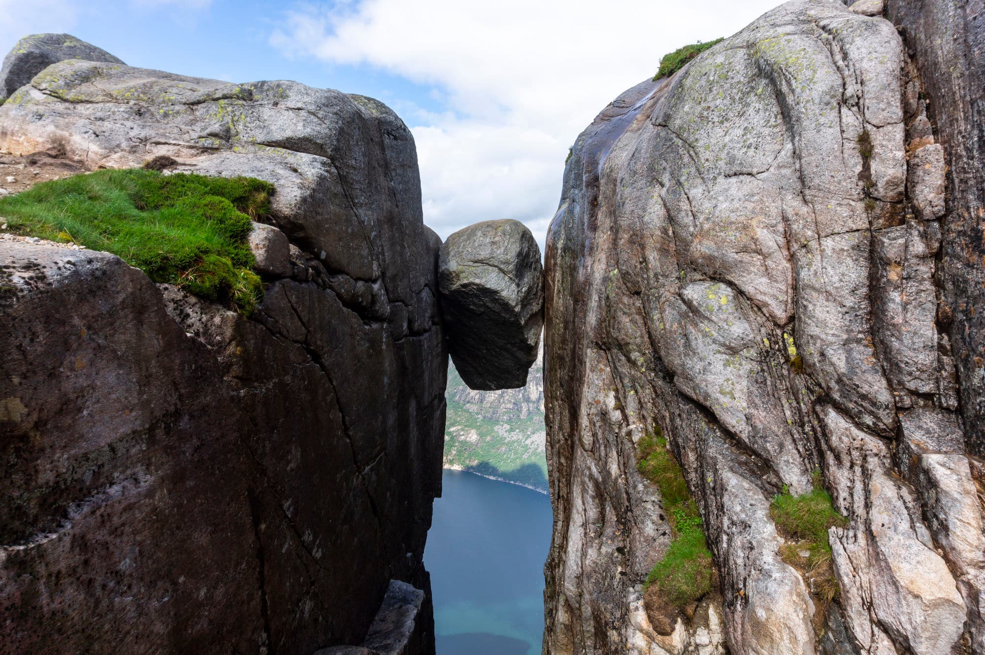 A large stone boulder wedged between two mountain sides. In the background, there is a fjord.