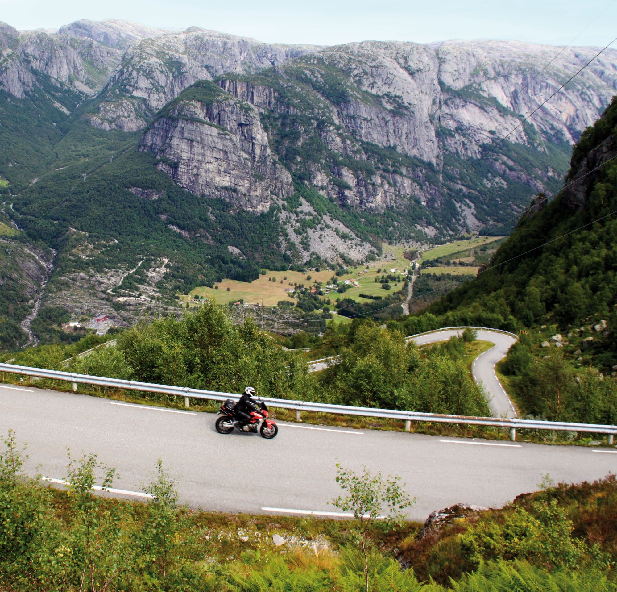 Person on motorbike driving on a road surrounded by large mountains, hairpin roads seen further down into a valley.
