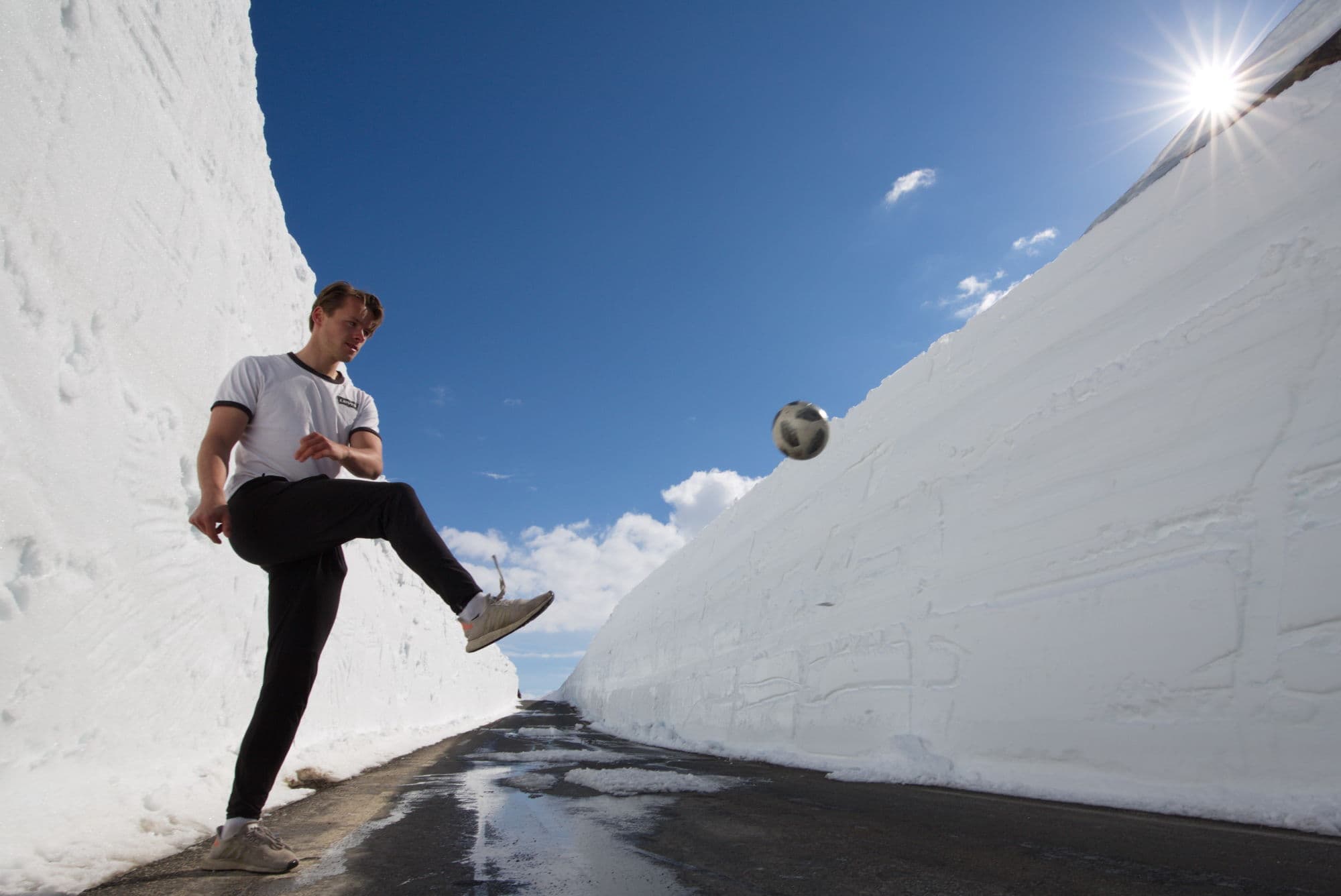 Man playing with a football on a road surrounded by high snowbanks on each side.