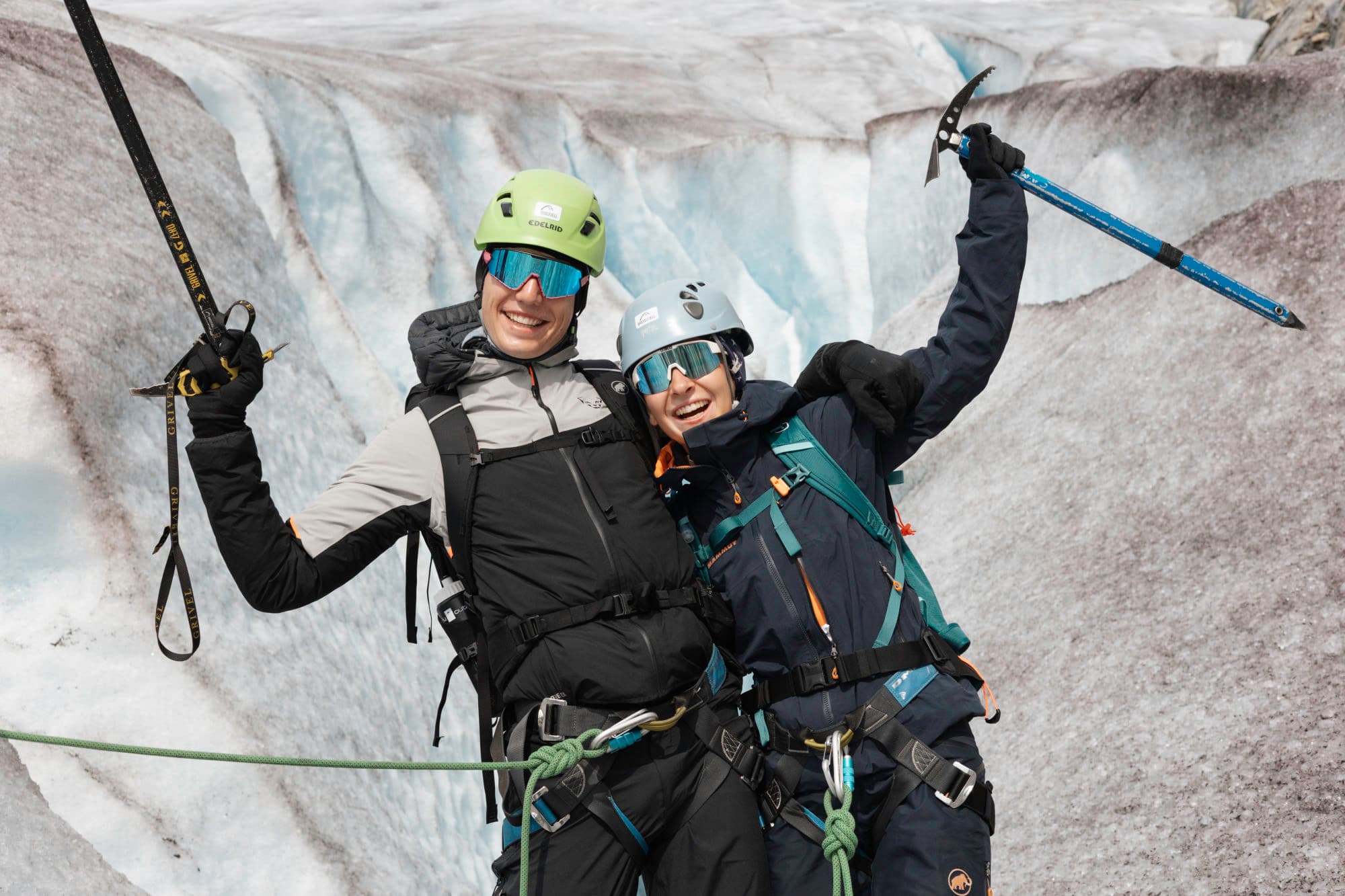 to personer smiler og holder redskapene sine i veret. de smiler til kamera, og står tett sammen. bak de er det en bre