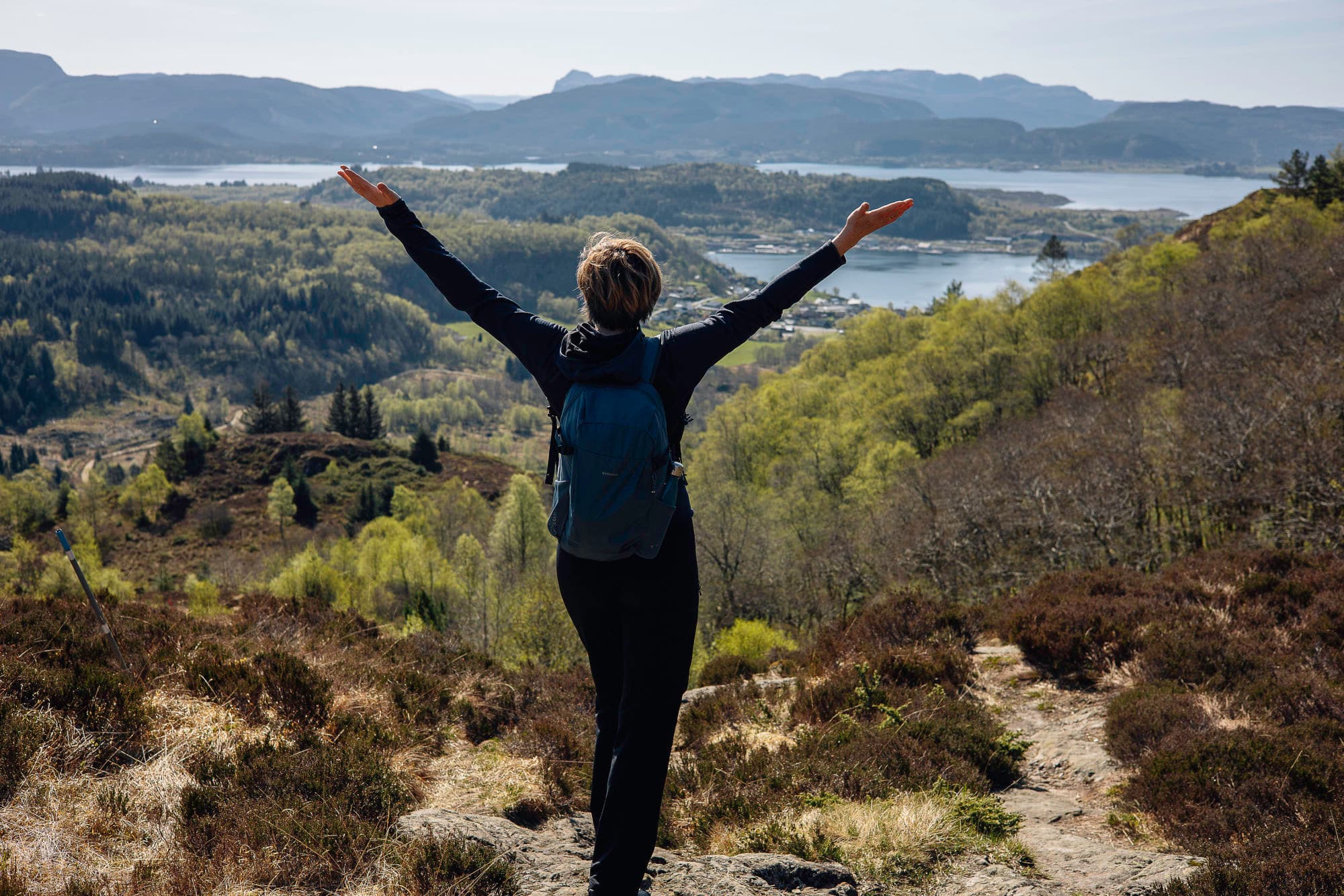 ei dame står med utstrakte hender og ser mot utsikten. det er trær, fjord, fjell, bygd bak ho