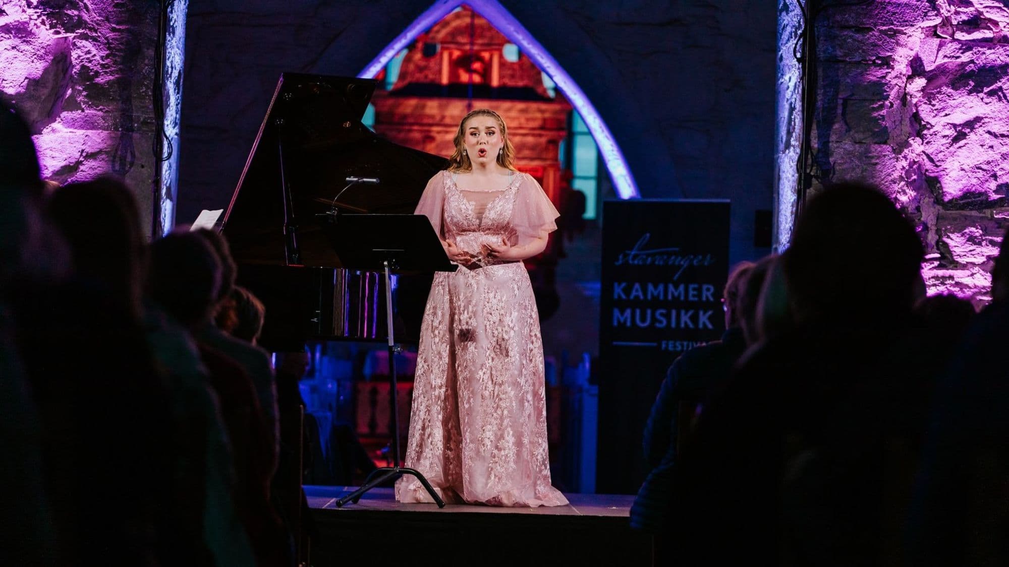Lady in pink ballroom dress on a stage singing. Behind her is a piano.