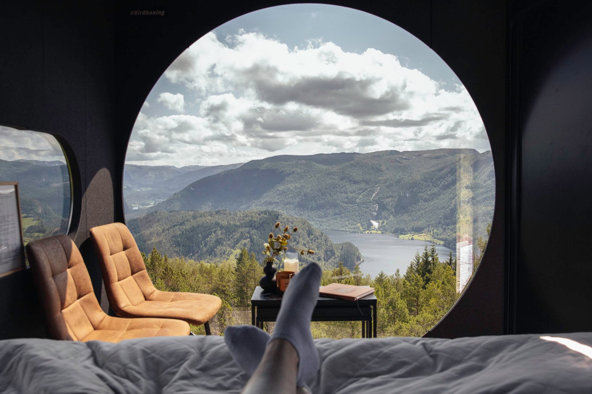 Eit par føte stikker i front av bilde. ho ligg på ein seng, og det er eit stort rundt vindauge foran ho. det er to stoler som står inntil vindauge, og eit bord