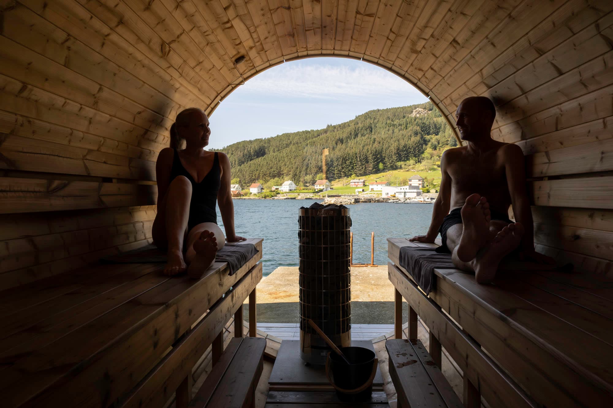 to personer, ein mann og ei dame sitter på kvar sin side i ein sauna. det er eit stort rundt vindauge som dei kan sjå ut. der ser du havet, og på andre sida er det mange hus, skog og åkre