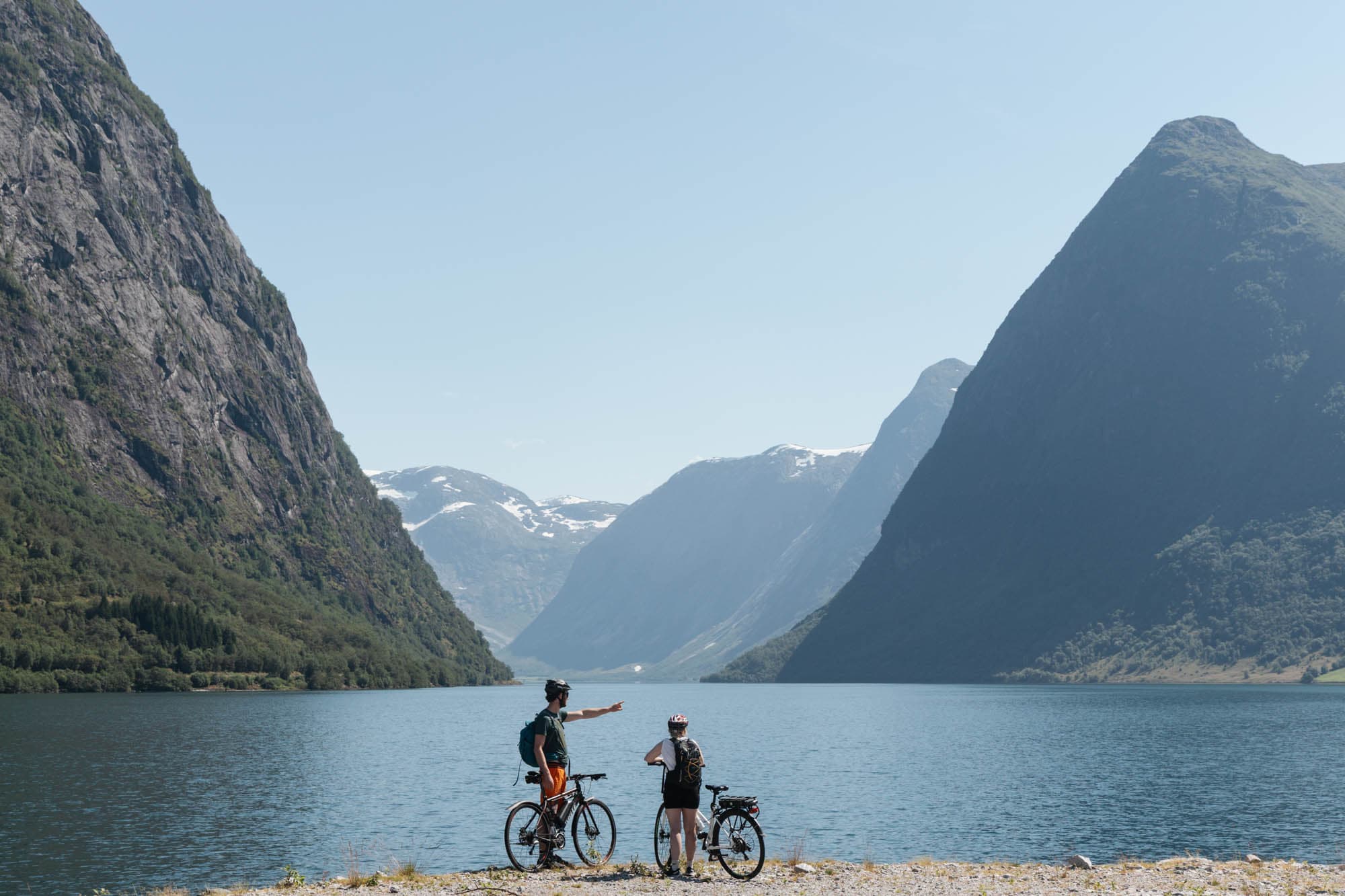 to personer står ved siden av sykkelen nærme eit vatn. guten peikar utover. det er høge fjell rundt dei