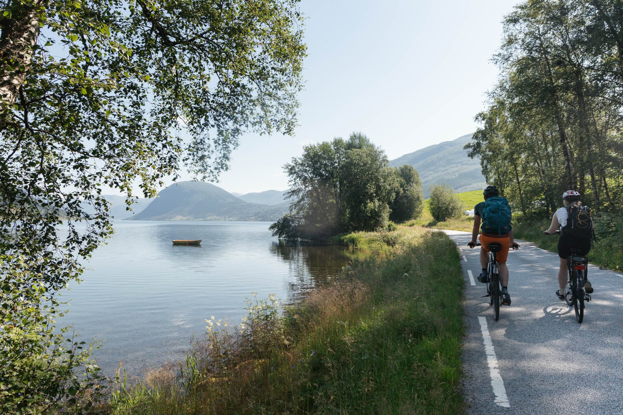 to personer sykler på en veg som ligg nærme eit vatn. på vatnet ligg ein båt, og det er skikkelig nasjonalromantisk stemning. det er sommar, blå himmel og grønne trær