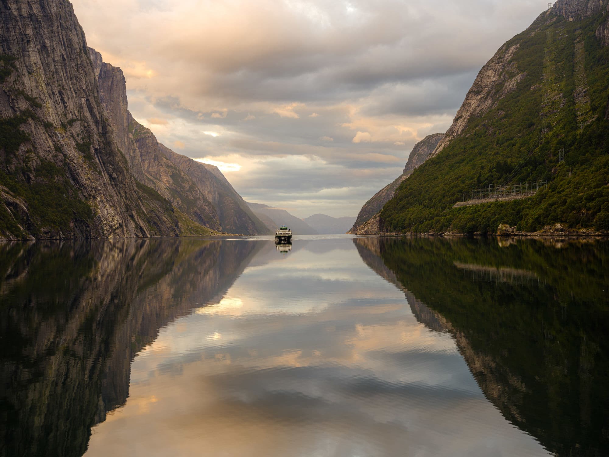 A boat is approaching ont the lysefjord, twe mountainsides on each sde reflects symmetrically into the fjord. The autumn colours are apparent