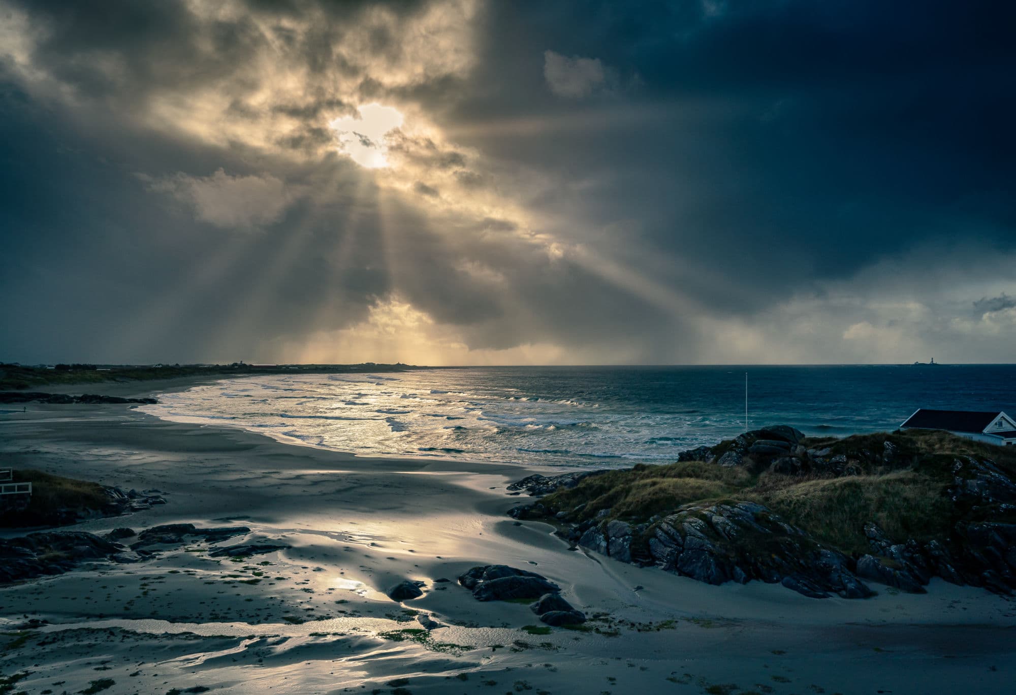 Solen skimtes gjennom et skydekke på en mørk høsthimmel. Strand, hav og svaberg i forgrunn.