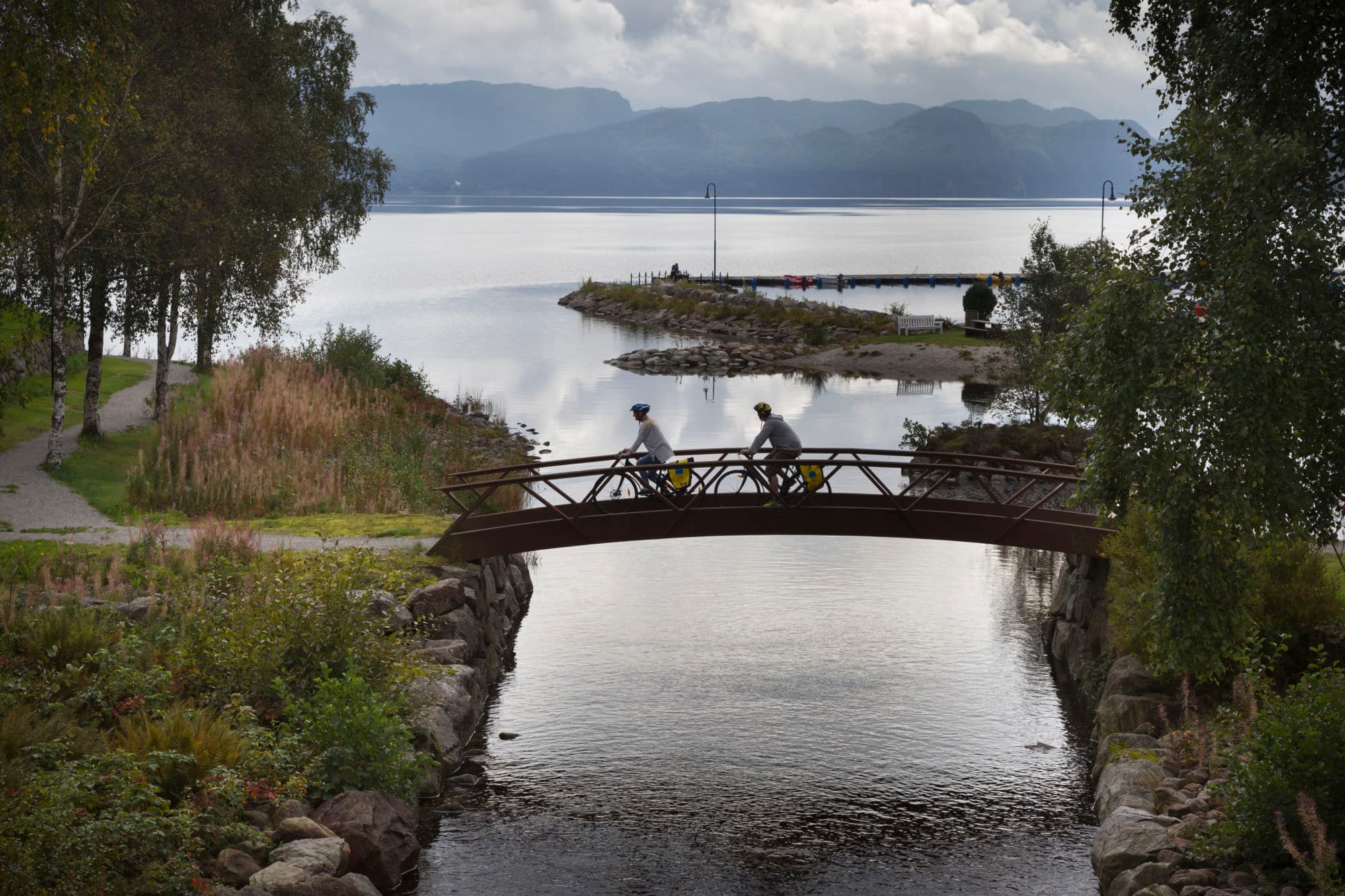 2 people cycling over a small footbridge over a lake