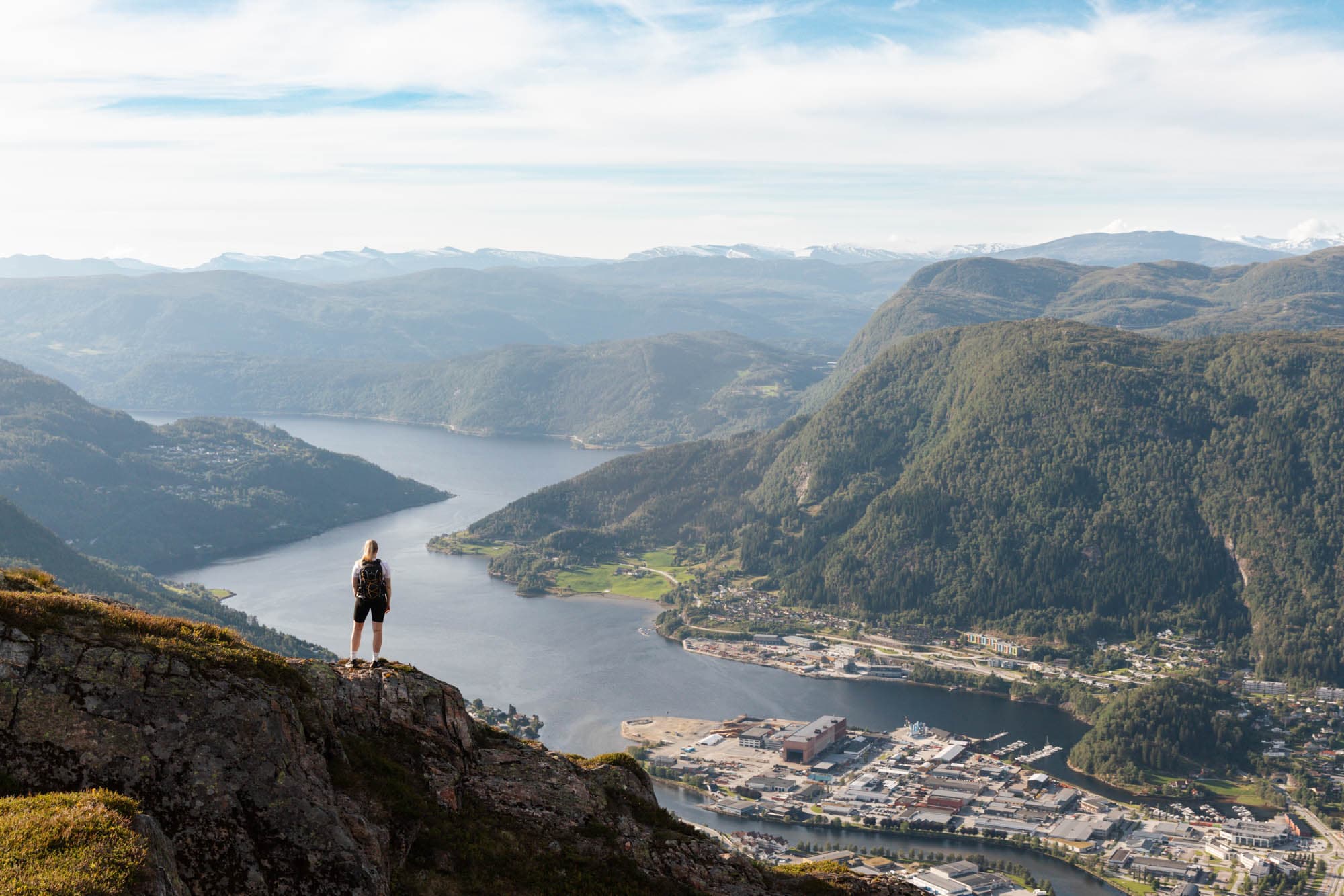 ein person går på eit fjell. utsikta bak er ein fjord med masse hus i enden. fjorden går i sikk-sakk utover med høge fjell på sidene