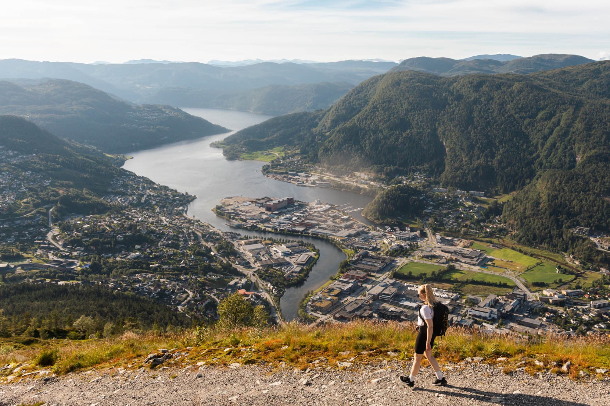 ein person går langs ein grusveg. bak ho er det utsikt over ein by som ligg i ein fjord. det er høge fjell mellom fjorden
