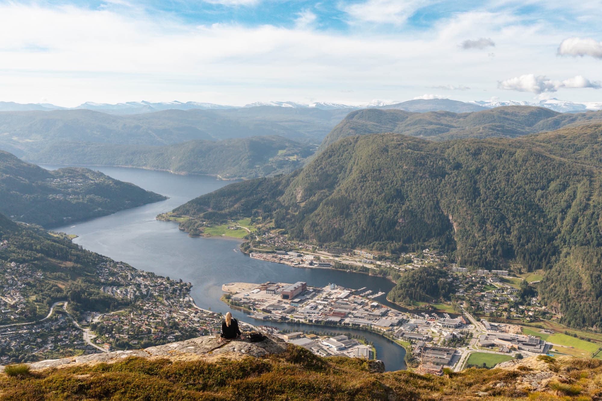 Ein person sitter på enden av eit fjell og ser ut over. utsikten er over ein by med masse hus, og ein fjord som går mellom høge fjell