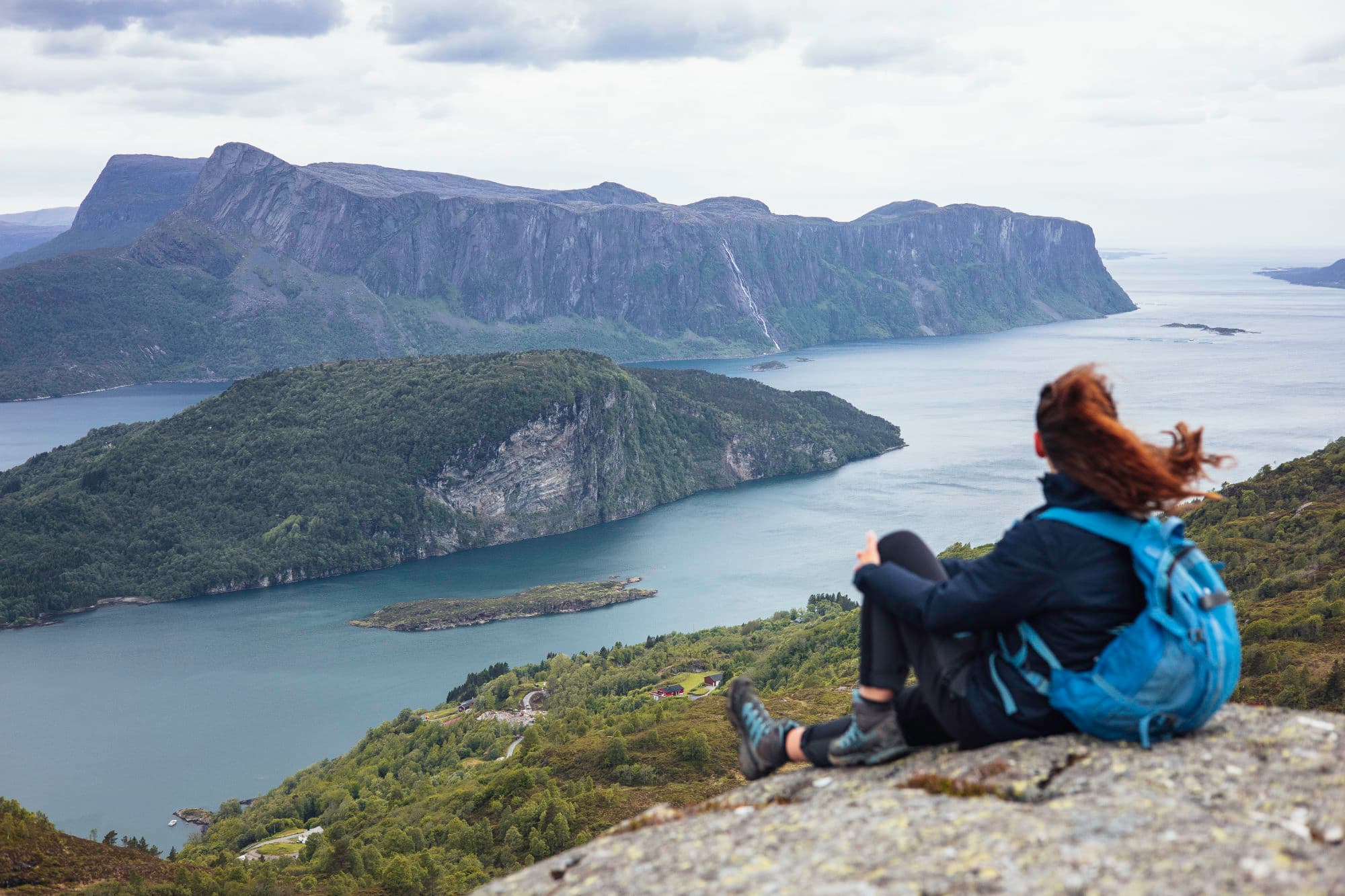 en person sitter oppå eit fjell. det er utsikt ut over eit stort fjell som stuper ned i ein fjord. fjellet er langt og bratt på sida. i midten av fjorden ligg det ei øy.