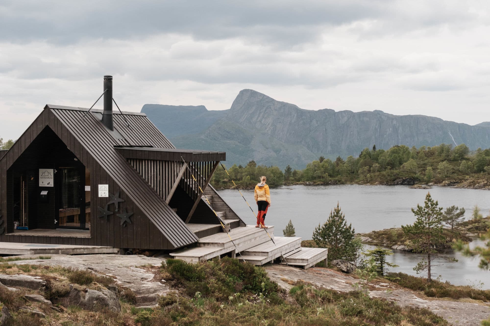 ein person står ved ei hytte, type dagsturhytte. ho ser utover det mektige landskapet som viser eit stort vatn i forgrunn, og eit stort fjell som stikker opp i bakgrunnen.