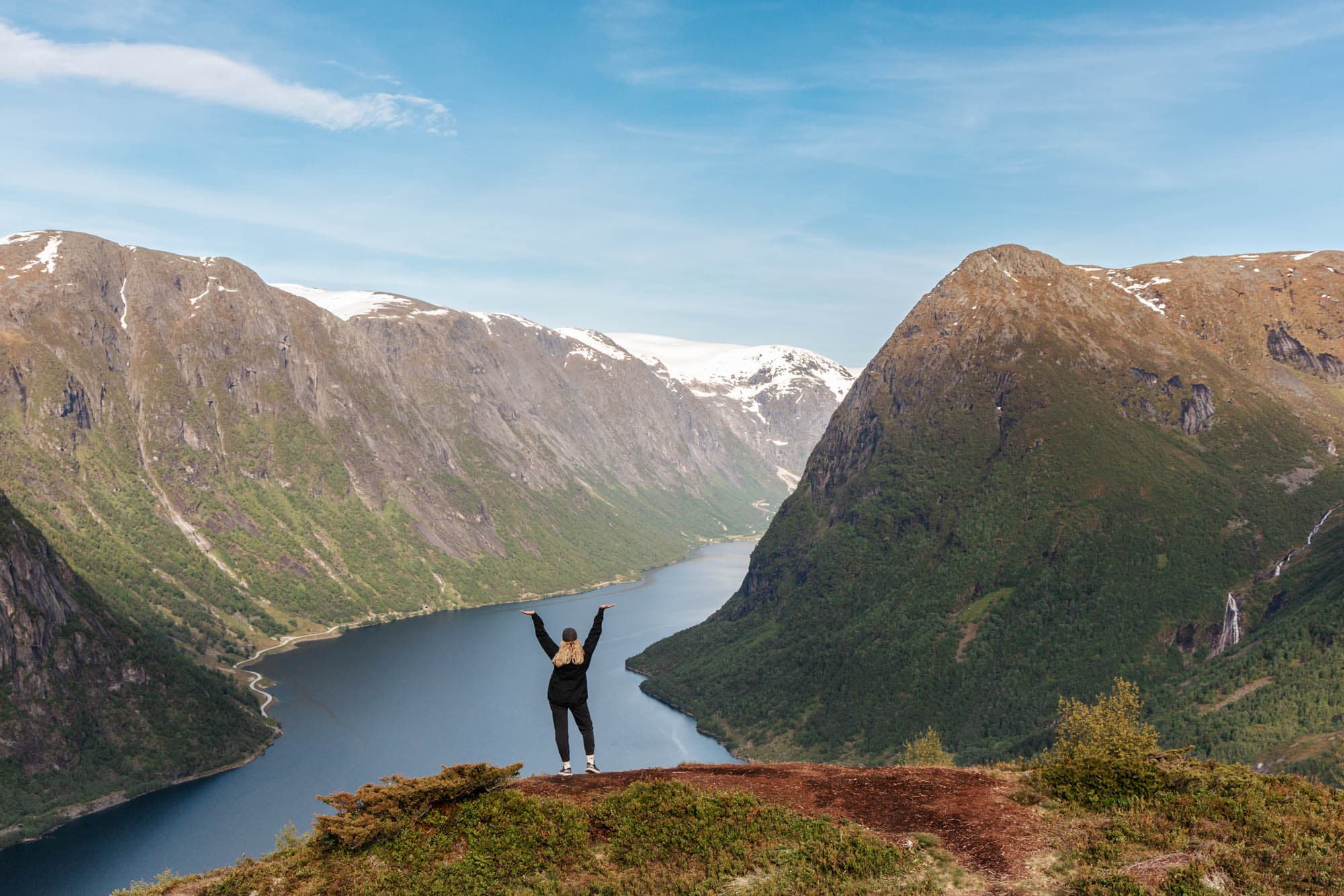 eit motiv med ein fjord og høge fjell med snøtoppar. det er grøne trer med eit hint av oransje. det er også grøne og oransje element i graset. i midten av bilde står ei jente og ser utover utsikten