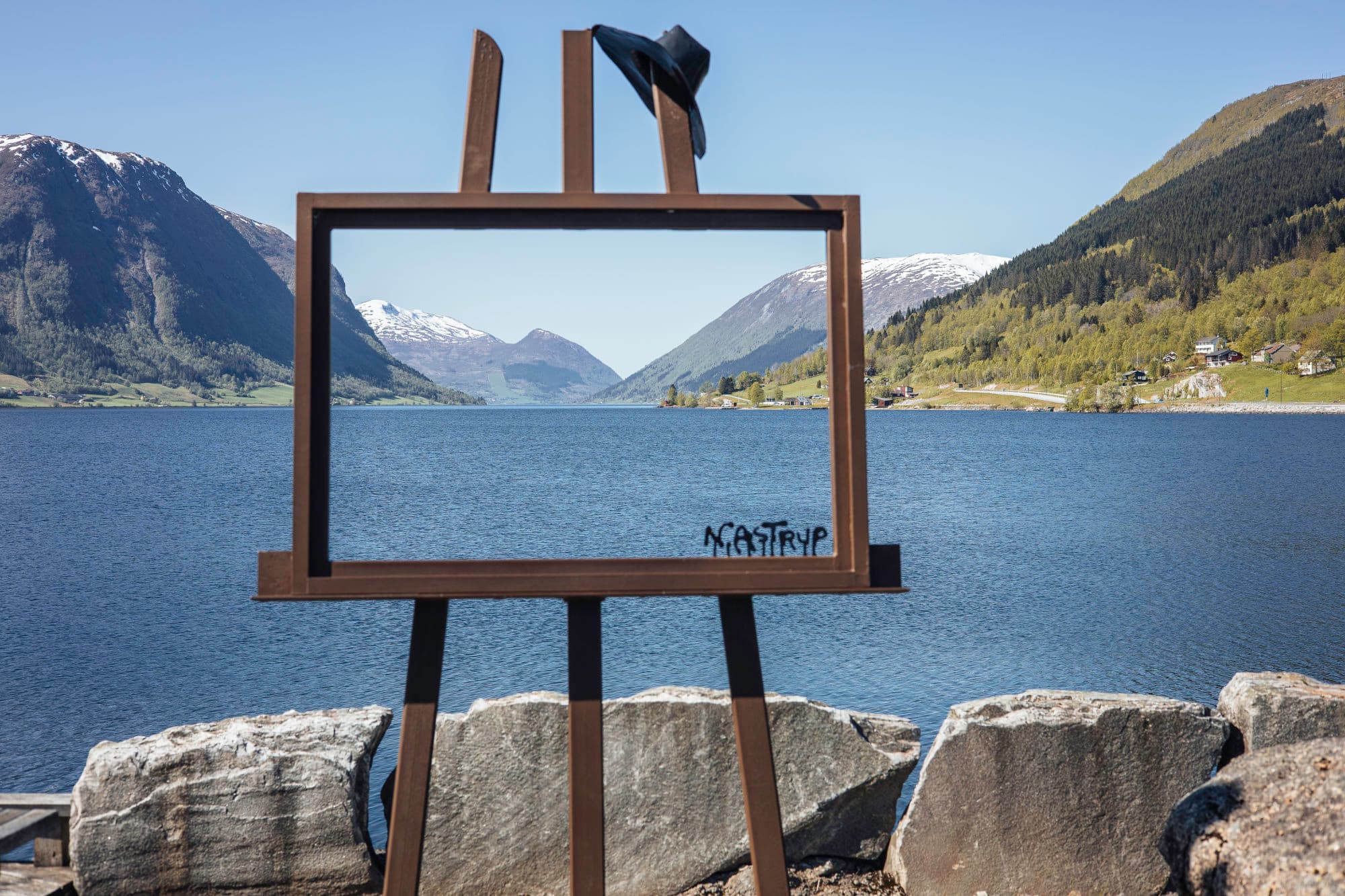 Ei ramme som står langs eit stort vatn. ramma er eit staffeli, inspirert av korleis nikolai astrup sitt var. bilde skal illustrere kor han fann motivene sine frå, jølster. det er høge fjell og snø på toppane rundt vatnet