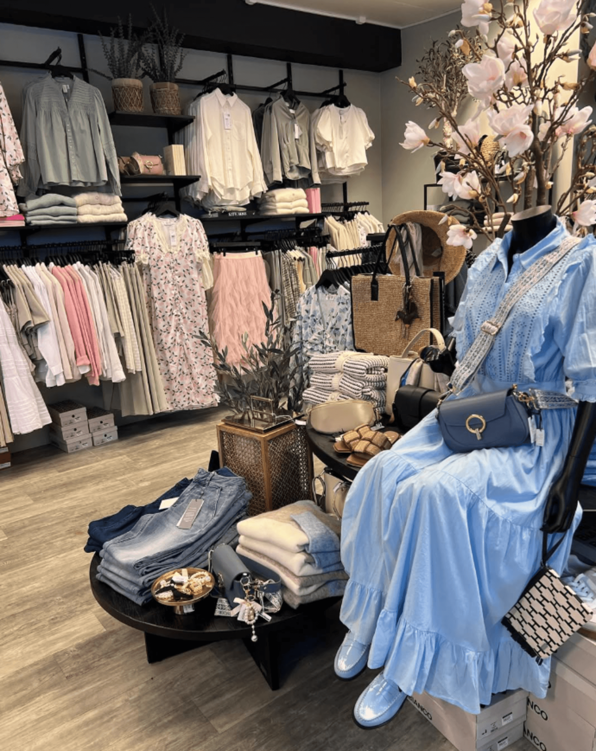 ein butikk med klede. i bilde ser du masse kle som henger fint opp på hyller. i front av bilde er eit bord med utstilling av bukser, vesker, ein blomst og ein manekeng av ein person som har på seg ein kjole, og veske