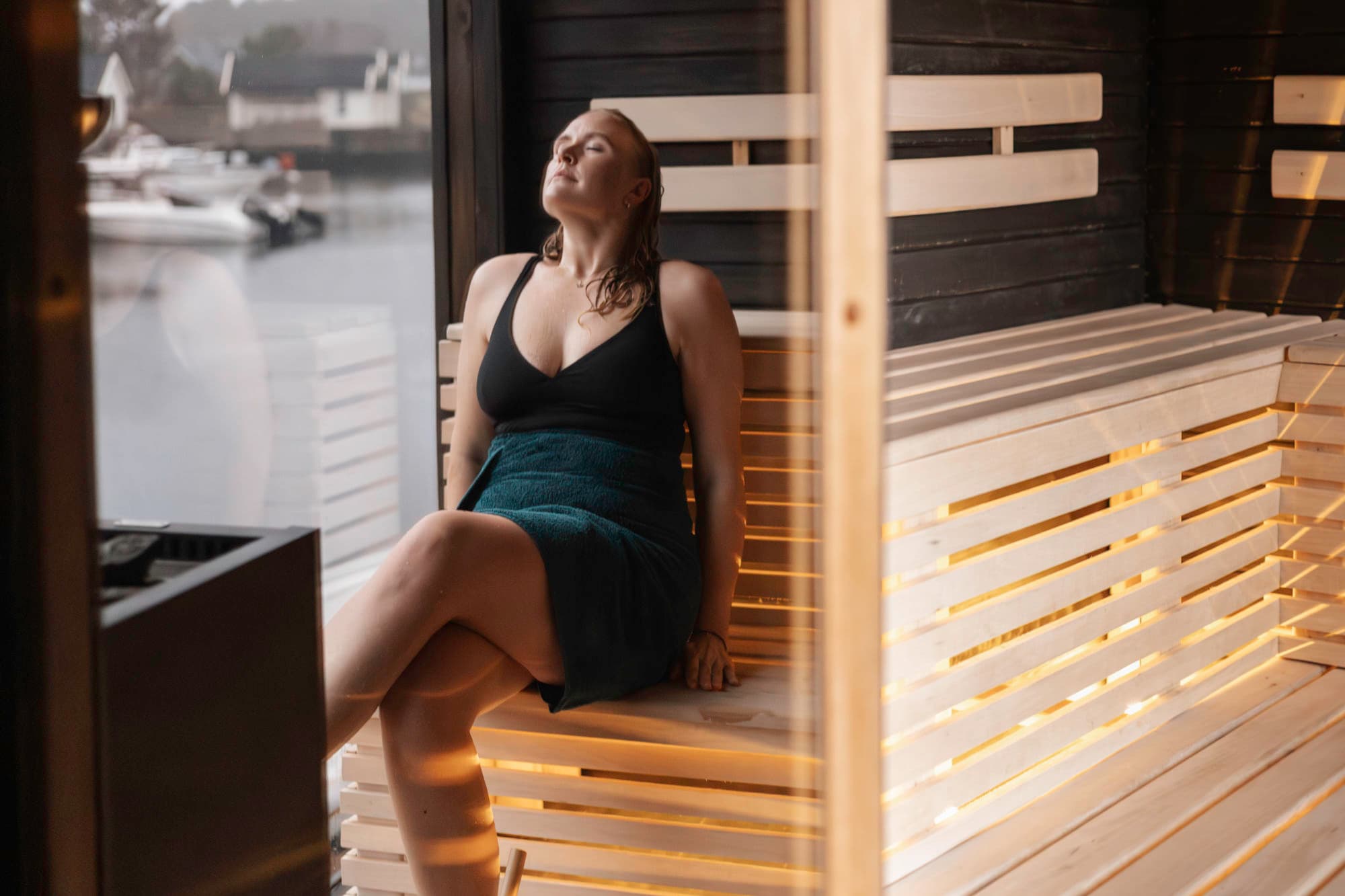 Ei jente sitter og nyter varmen i ei badstue. ho har augene lukka, og ho ser oppover. ho har hendene på sida av seg. ho har beina i kryss, har på seg svart badedrakt og grønt handklede