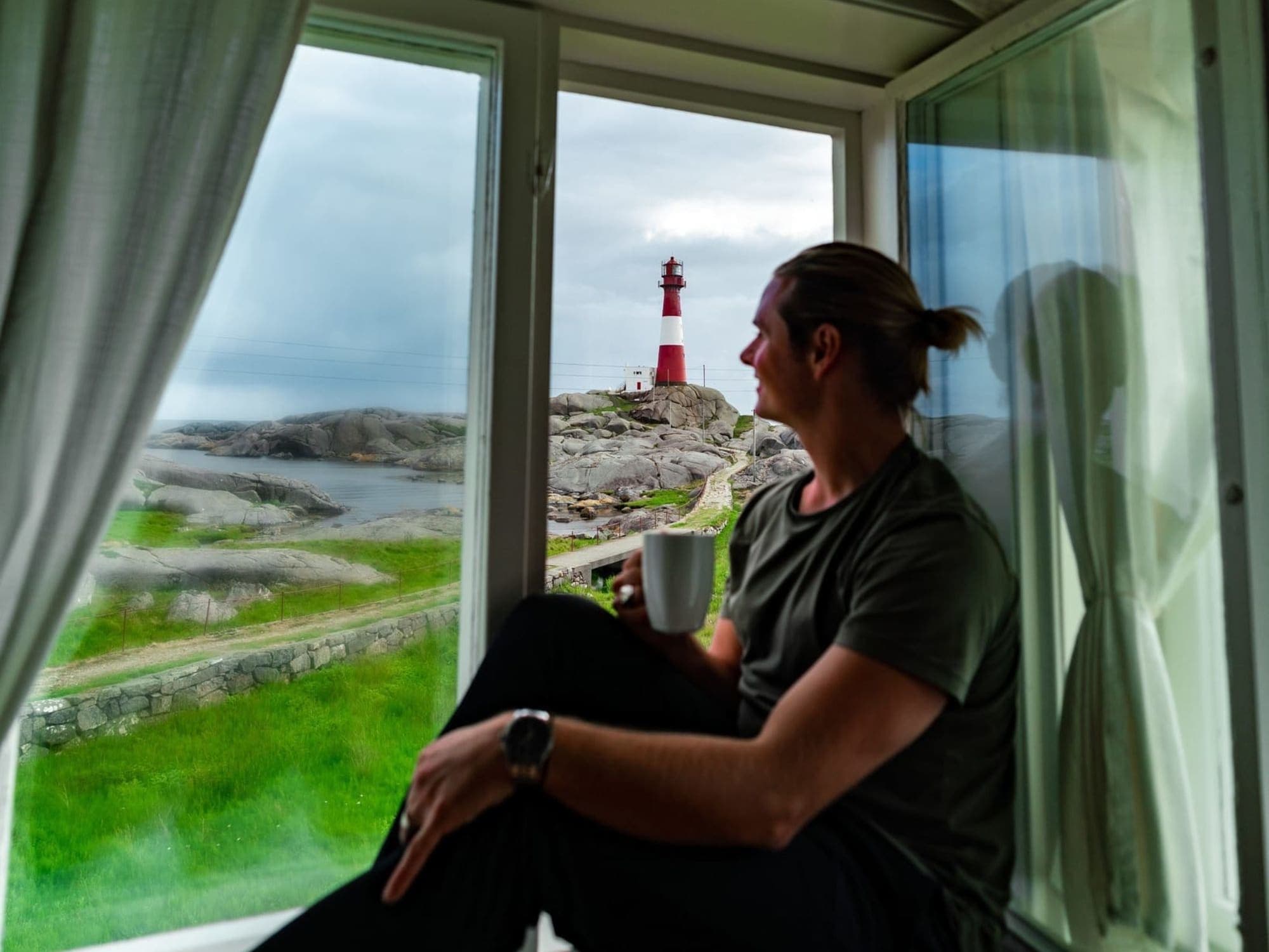 Person sitting on window sill looking out an open window with view of the ocean and lighthouse