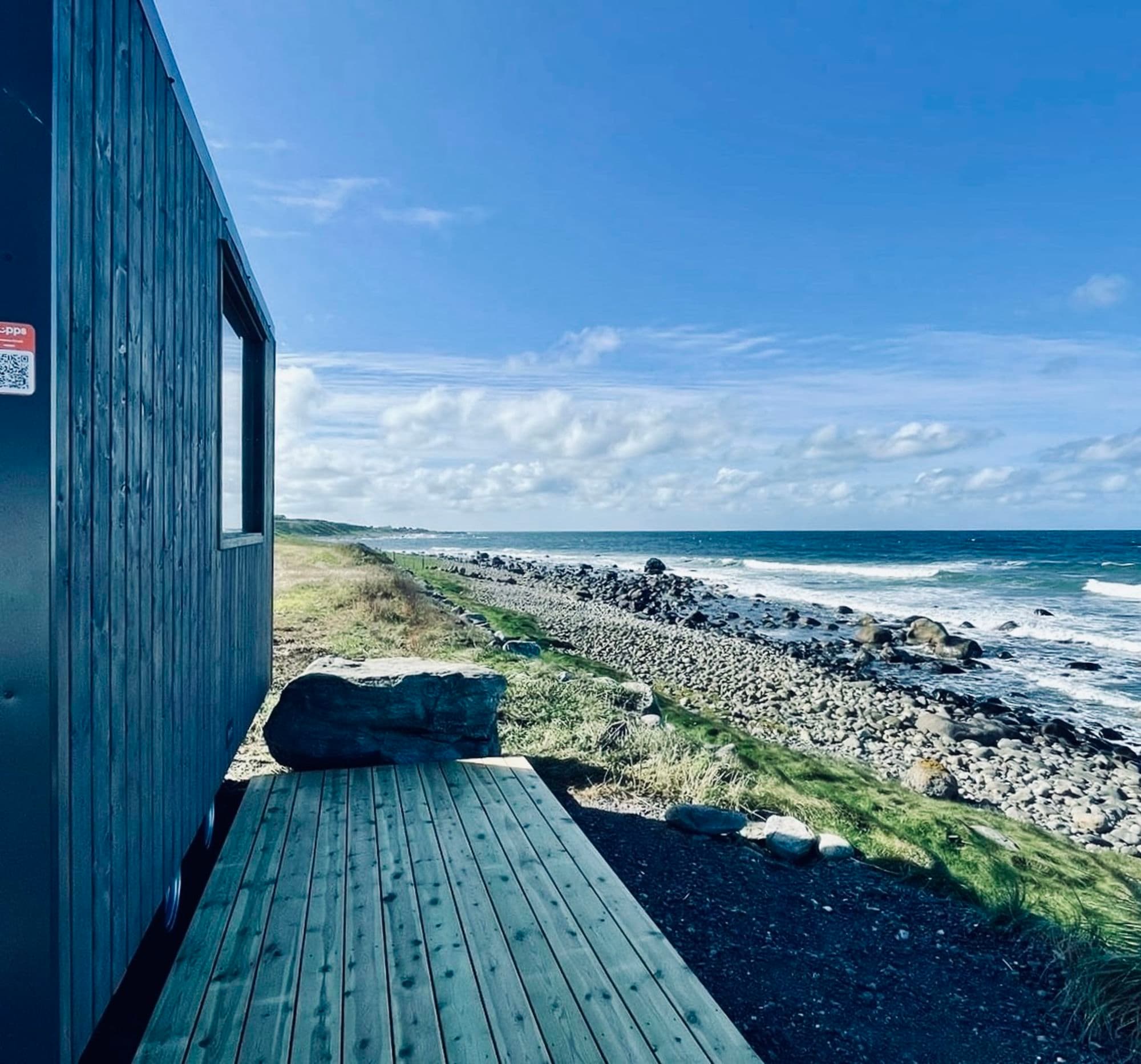 BookSauna the deck of a sauna and the wooden wall of the sauna facing the sea with round rocks.