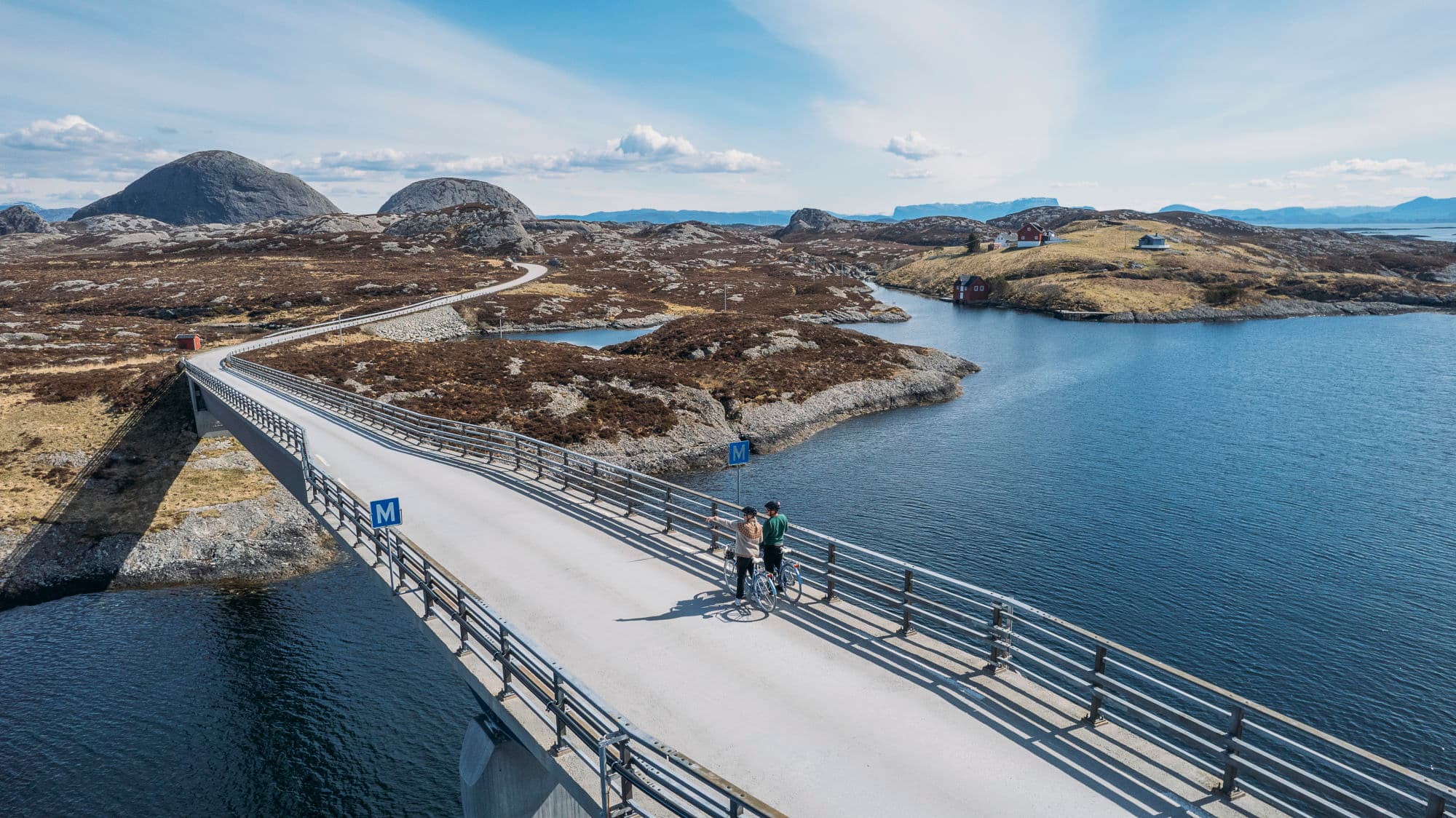 to personer står på sykkelen sin på ein bru. det er holmer og skjær rundt dei. det er blått hav og blå himmel
