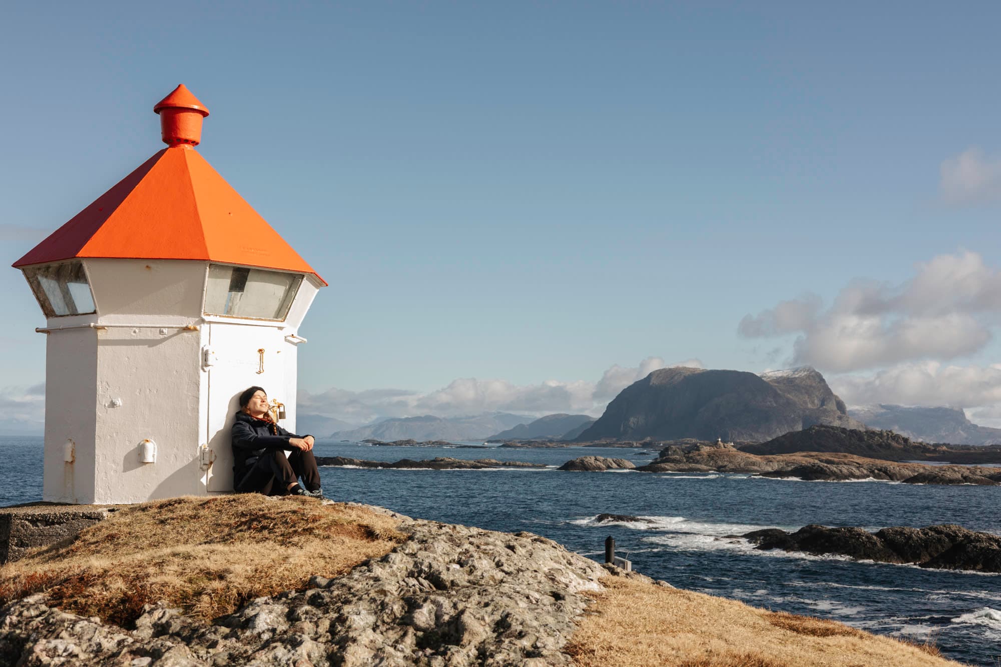ei jente sitter ved ei fyrlykt og nyter sola. det er blått ope hav rundt ho med holmar og skjer