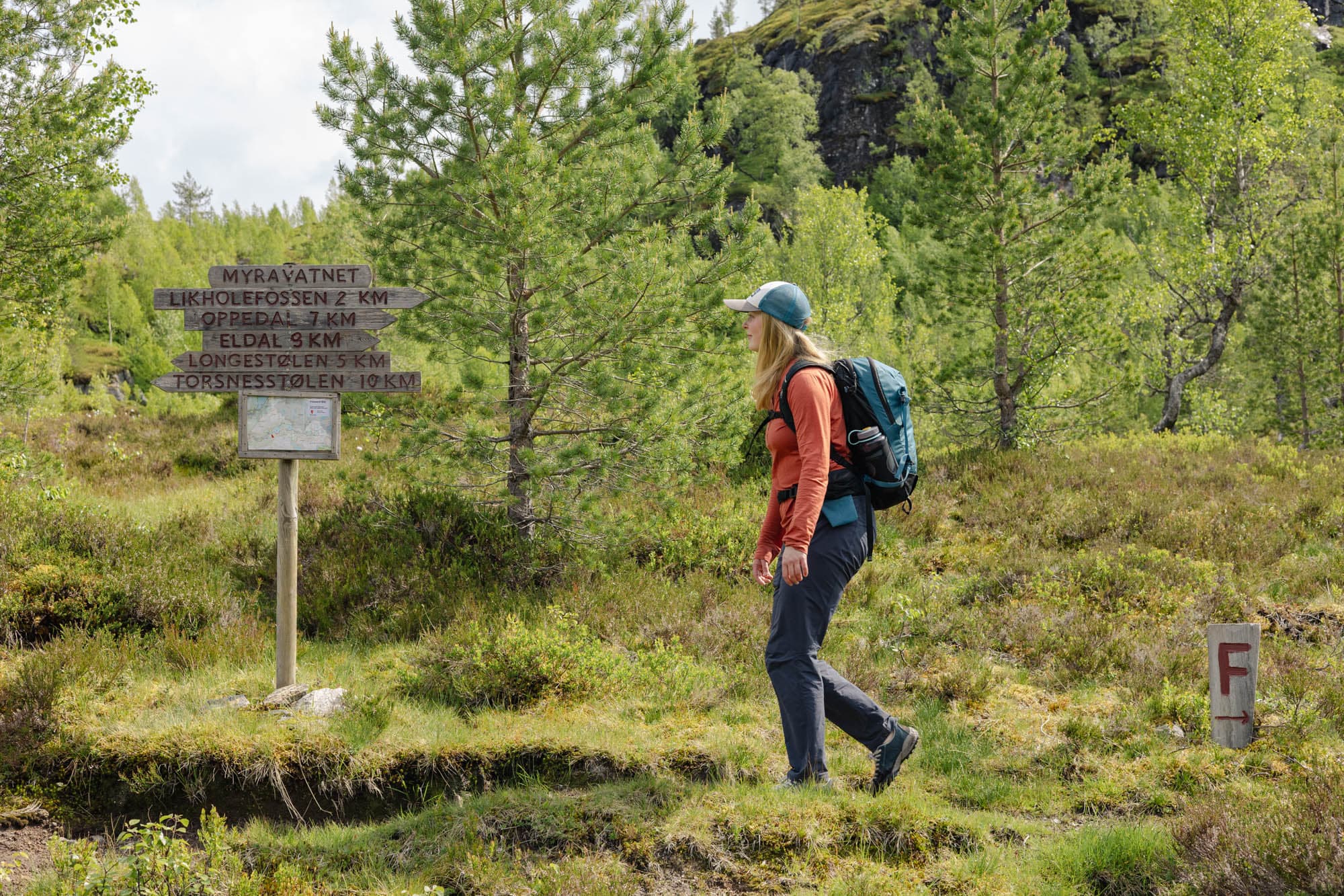 Ei jente går forbi fleire skilt som står i naturen. ho har på seg turklede, caps og sekk. det er grøne element rundt ho.