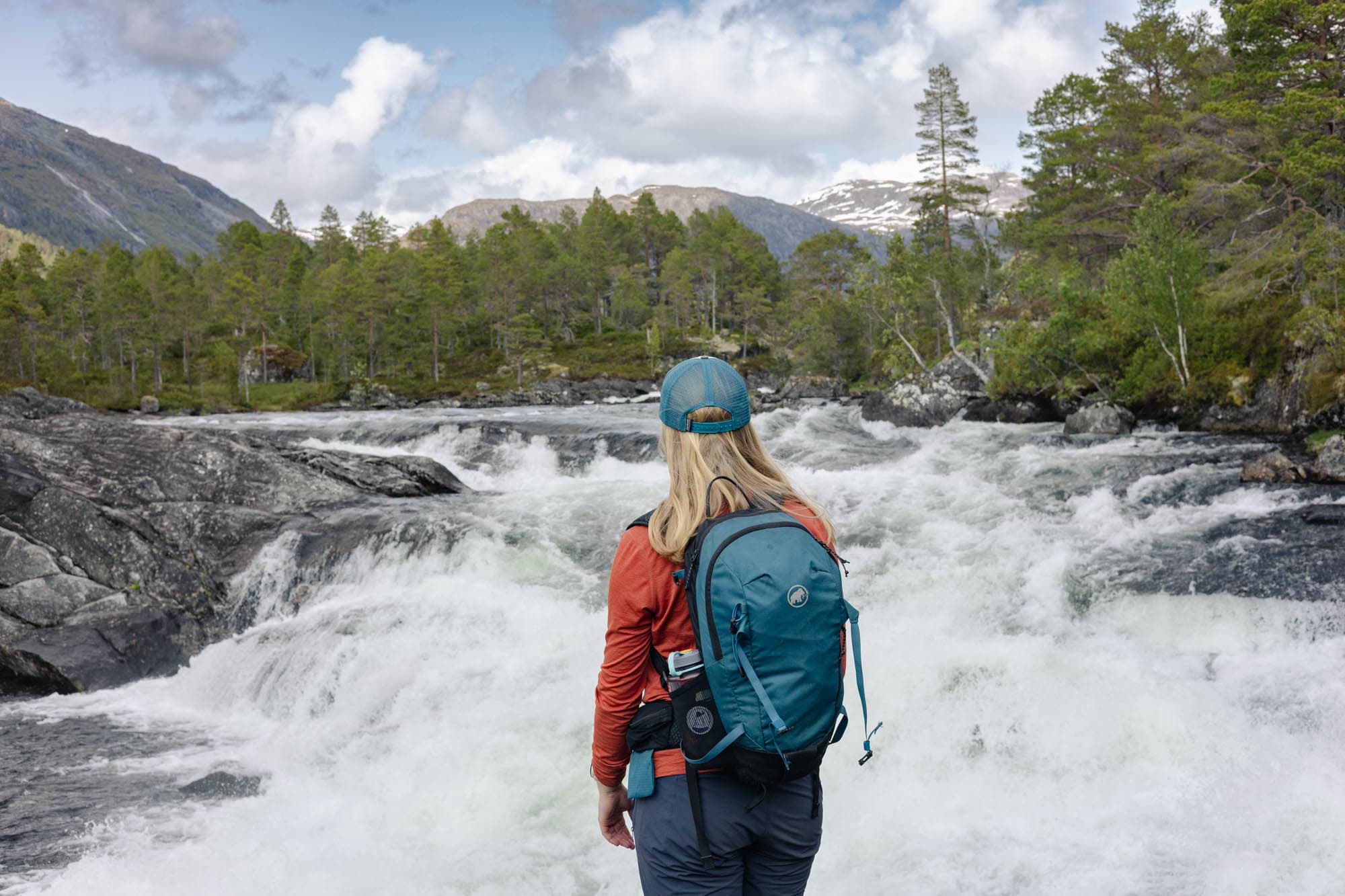 Ei jente står og ser på ein foss som går forbi ho. det er trær i bakgrunnen. jenta har på seg turklede, sekk og caps