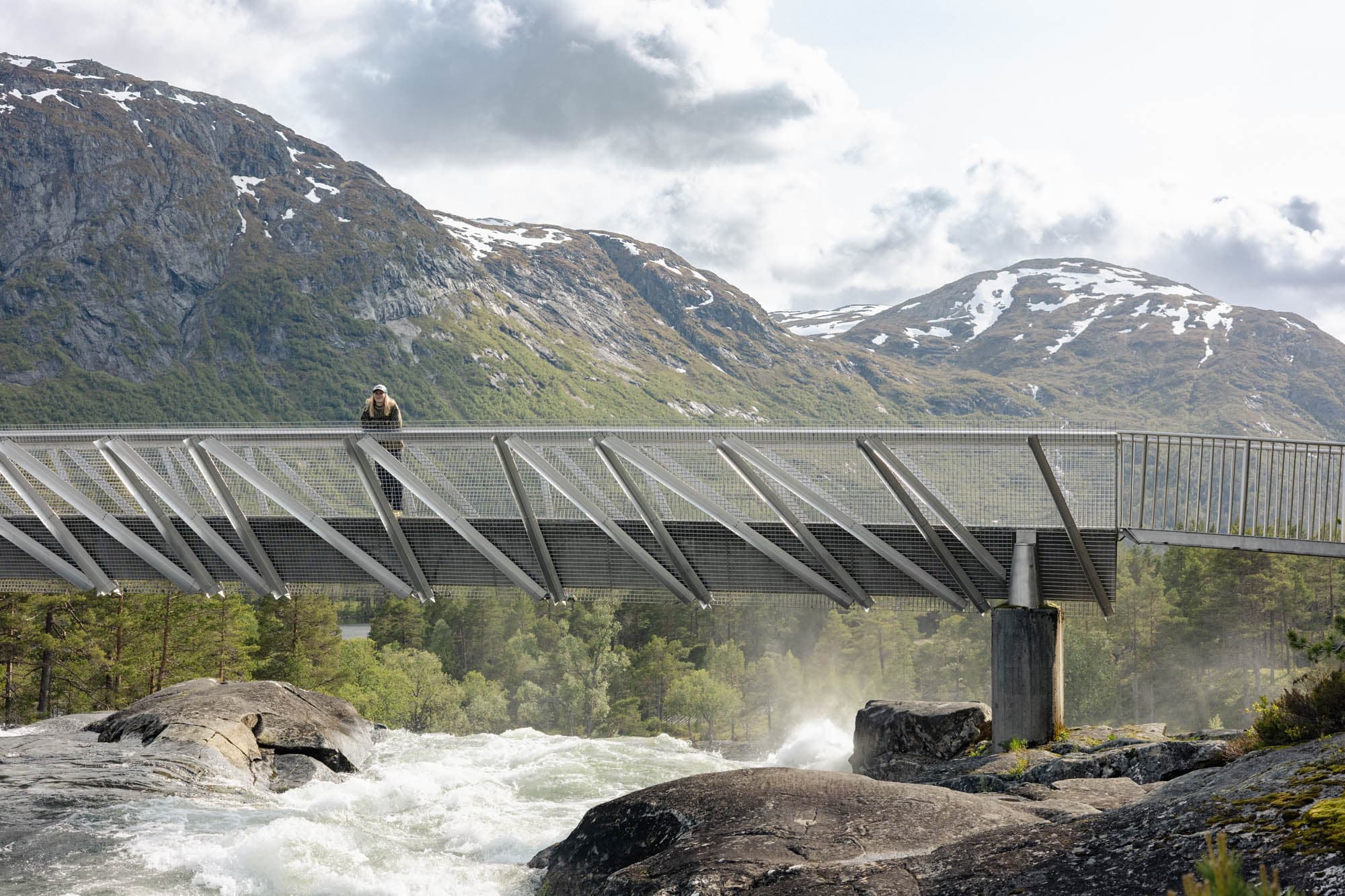 Ei jente står på ein bru som går over ein foss. ho ser bort mot kamera og smiler. fossen bruser. bak ho er mektige fjell med snø på toppen