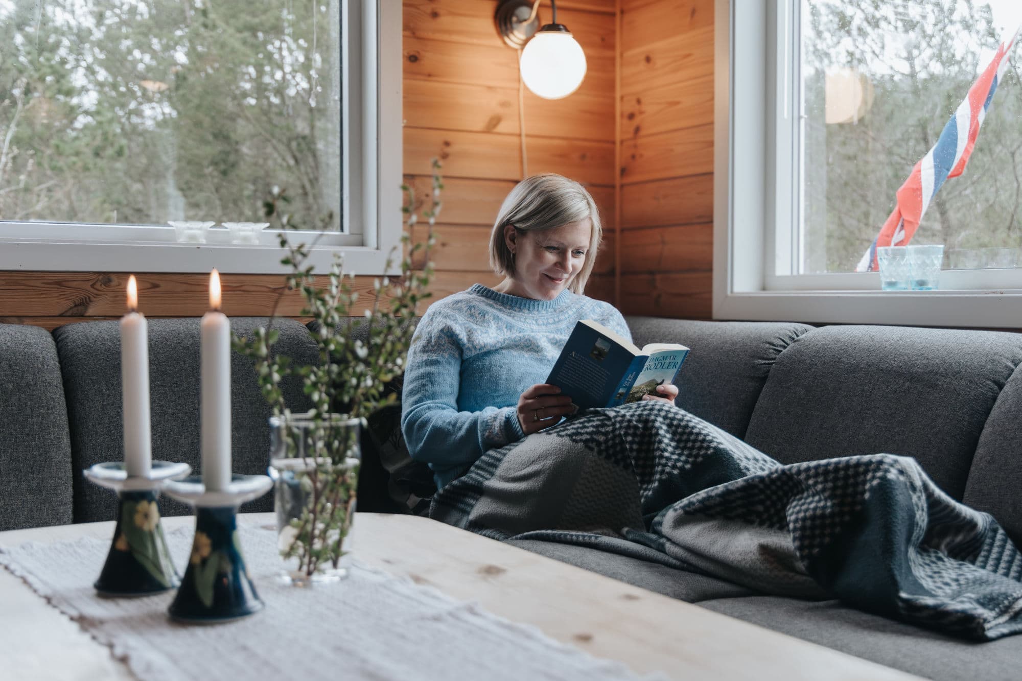 Ei dame sitt i ein sofa med pledd på og les ein bok. det er store vindauge ved siden av ho, og i eine vindauge heng det ein vimpel ute. ho har eit bord foran seg med levande lys og blomar.