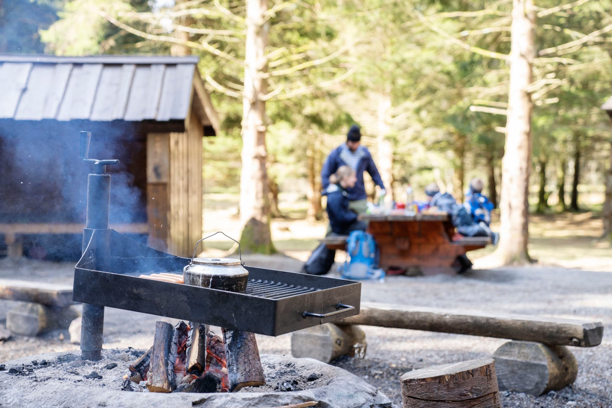 Barbecue area with sausages grilling and coffee brewing, family in the background